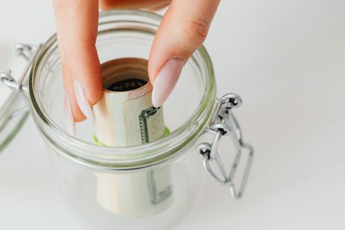 Free Close-up of hand placing rolled currency into a clear glass jar, symbolizing savings. Stock Photo