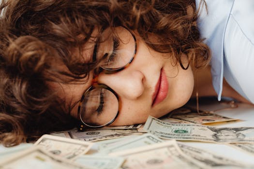 Close-up of a sleeping woman with curly hair and eyeglasses, resting on a pile of cash.