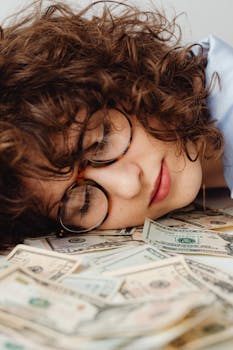 A woman with curly hair and glasses rests on a pile of US dollar bills.
