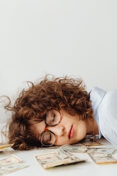 Caucasian woman with curly hair and eyeglasses sleeps on US dollar bills, illustrating finance and wealth concepts.