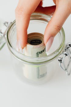 Close-up of manicured hand placing rolled-up cash into a clear glass jar for savings.