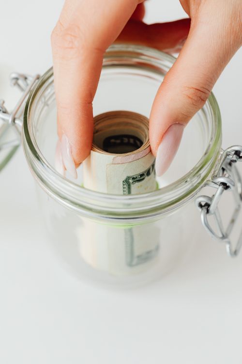Free Close-up of manicured hand placing rolled-up cash into a clear glass jar for savings. Stock Photo