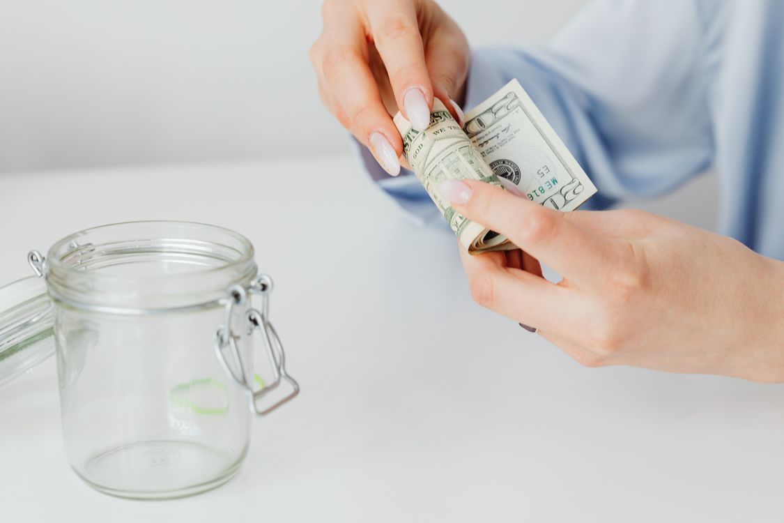 Free Woman rolling dollar bills beside a glass jar, symbolizing savings or financial planning. Stock Photo