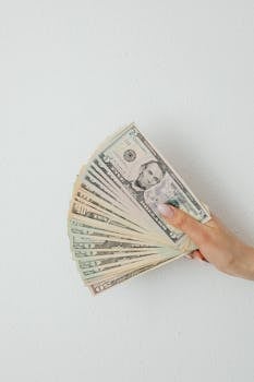 Photo by Karola G Close-up of a hand holding a fan of US dollar bills against a white surface.