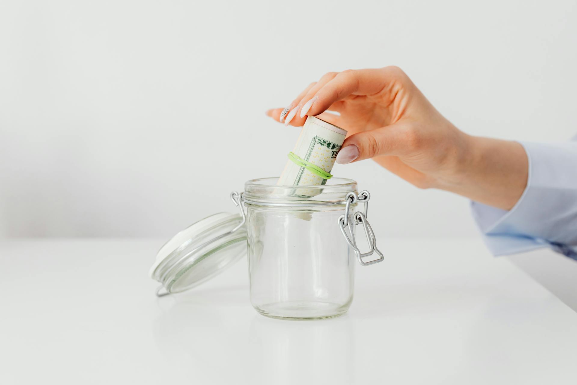 A close-up of a woman's hand putting rolled US dollar bills into a glass jar, symbolizing saving and budgeting.