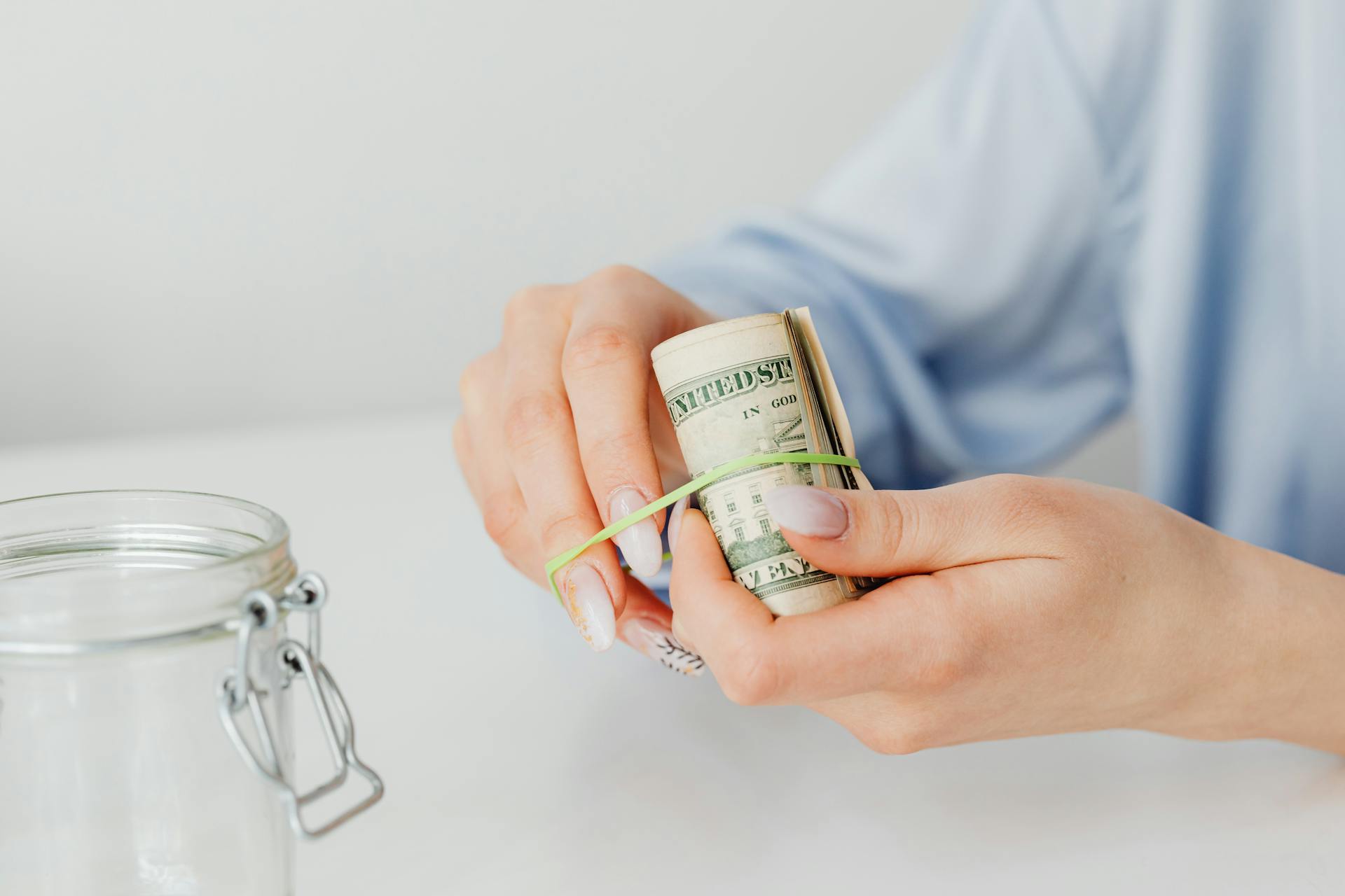 Close-up of hands securing rolled US dollar bills with rubber band beside a glass jar.