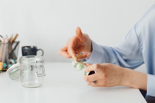 Close-up of hands rolling US dollars with a rubber band near an empty glass jar.