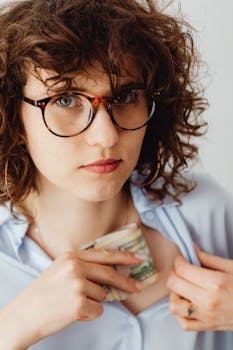 Woman wearing glasses tucks money into her shirt, expressing focus and caution.