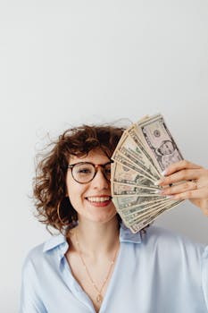 Smiling woman with curly hair holding a fan of dollar bills indoors.