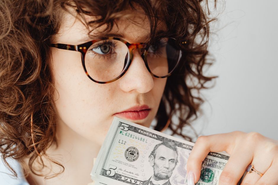 Photo by www.kaboompics.com on Pexels Close-up of a woman holding a US five dollar bill, wearing glasses and looking thoughtful.