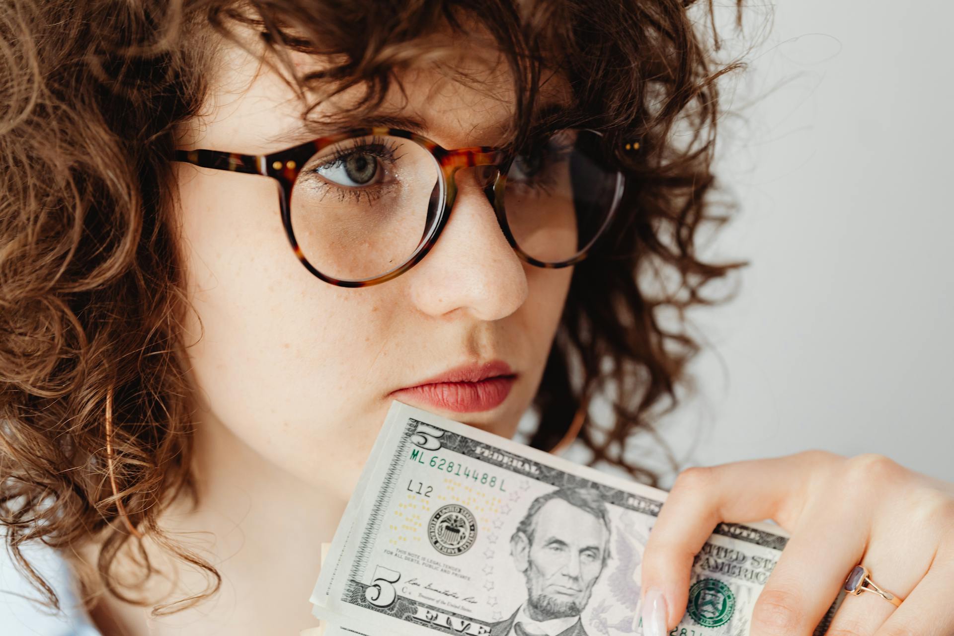 Close-up of a woman holding a US five dollar bill, wearing glasses and looking thoughtful.