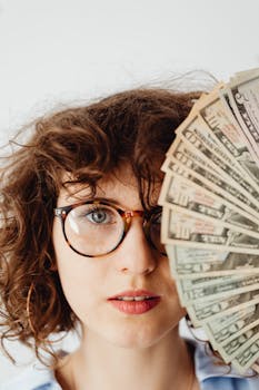 Portrait of a woman with eyeglasses holding a fan of US dollar bills close to her face.