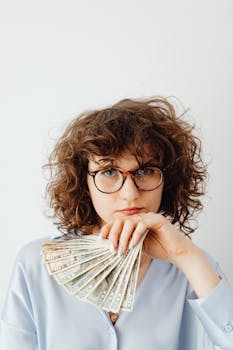Caucasian woman with curly hair and glasses holding US dollars confidently indoors.