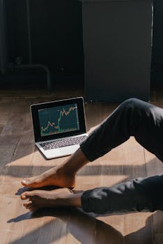 A person sits indoors with a laptop displaying stock market data on wooden flooring.