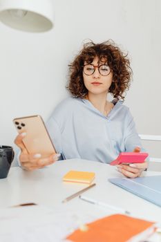 Woman with curly hair using smartphone at desk with notes and eyeglasses, indoors.