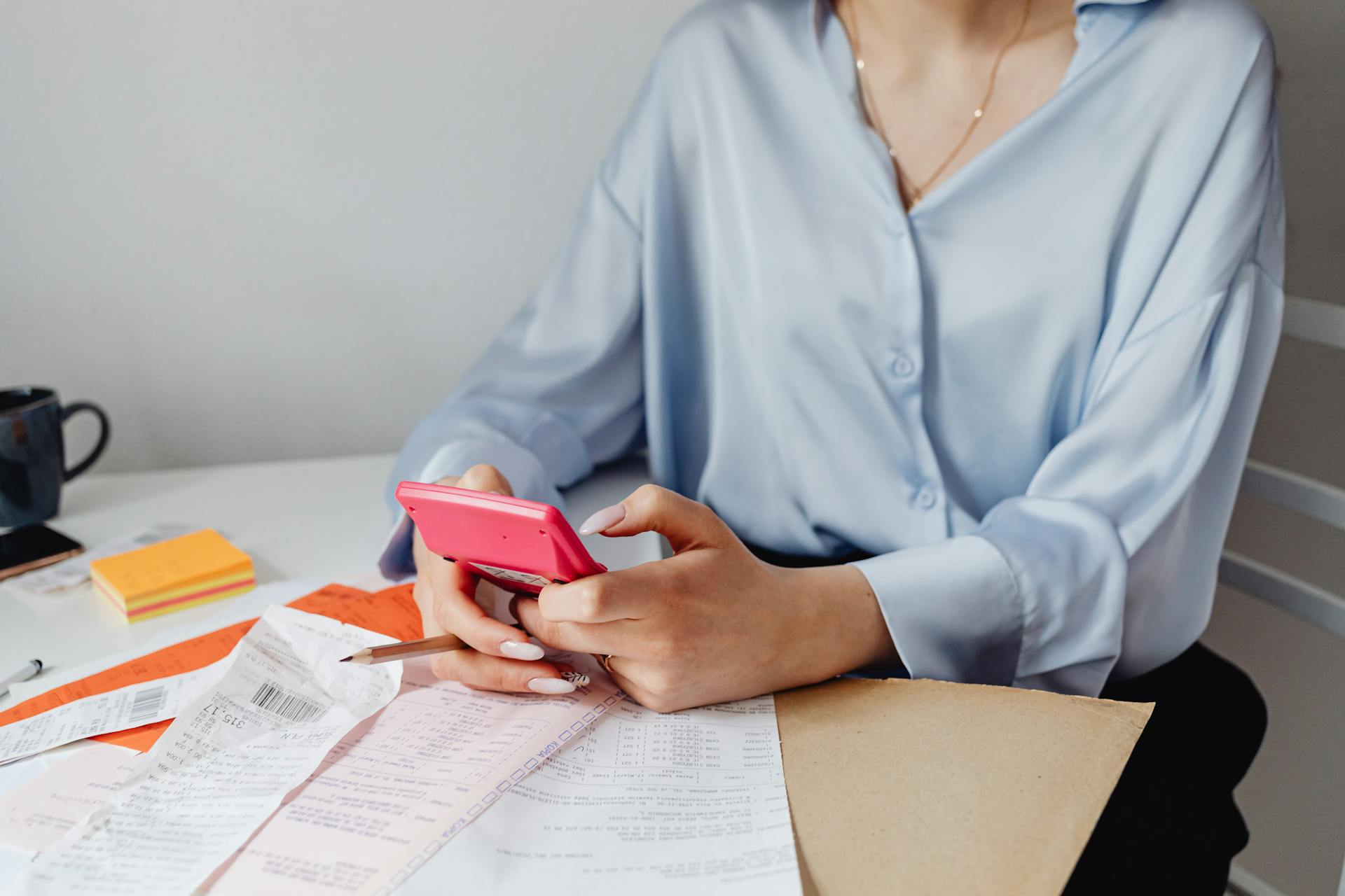 A woman using a pink calculator surrounded by bills and receipts at a desk.