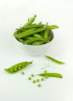 A white ceramic bowl filled with fresh green peas and pea pods on a light background.