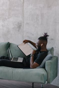 Bearded man working on a laptop while reclining on a sofa, analyzing data on a clipboard.