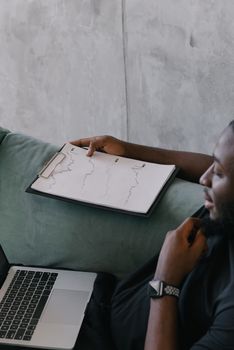Focused businessman in black shirt reviews financial graphs on clipboard beside laptop. Concrete wall background.