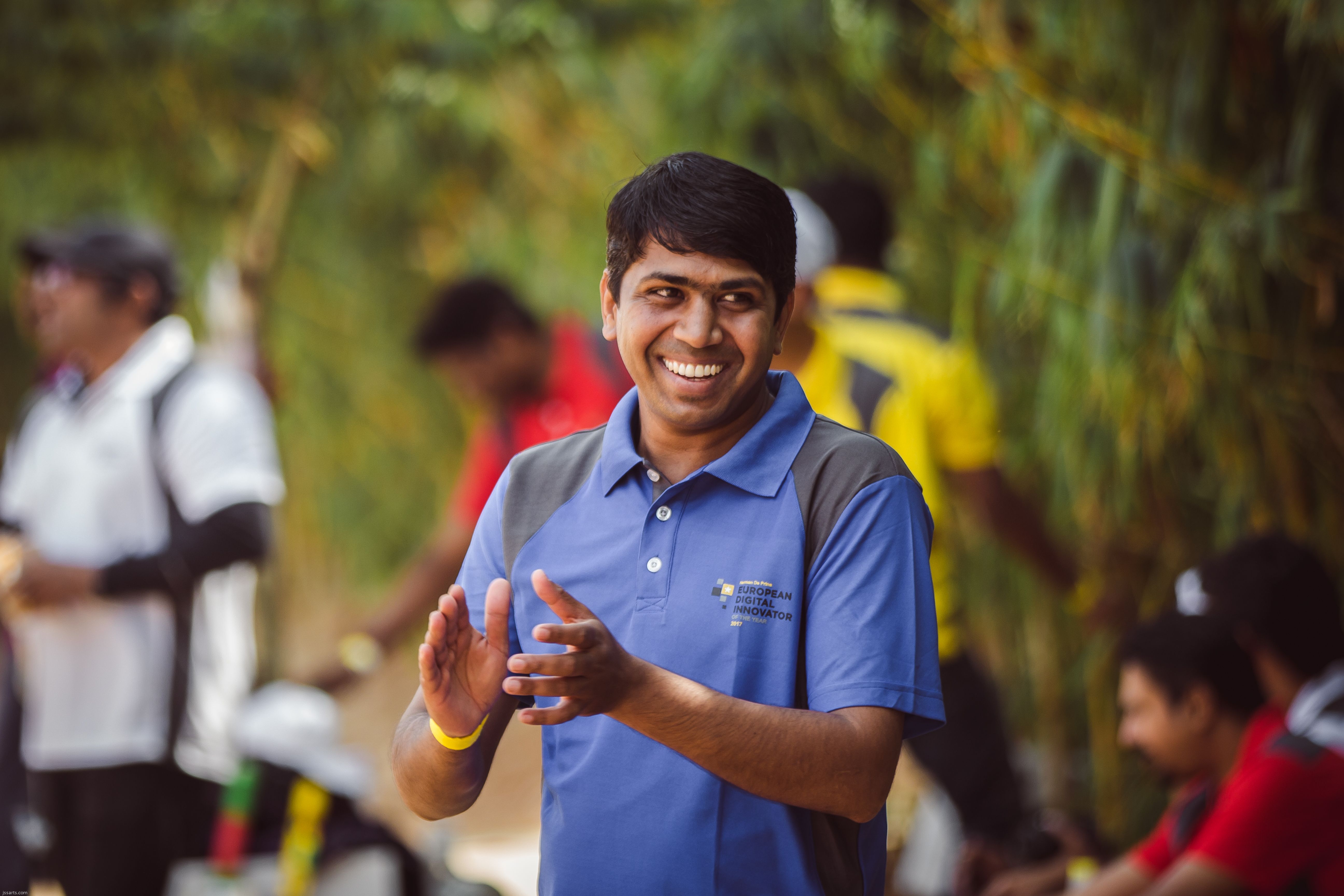 A cheerful man claps and smiles outdoors in a casual polo shirt, surrounded by people enjoying a sunny day.