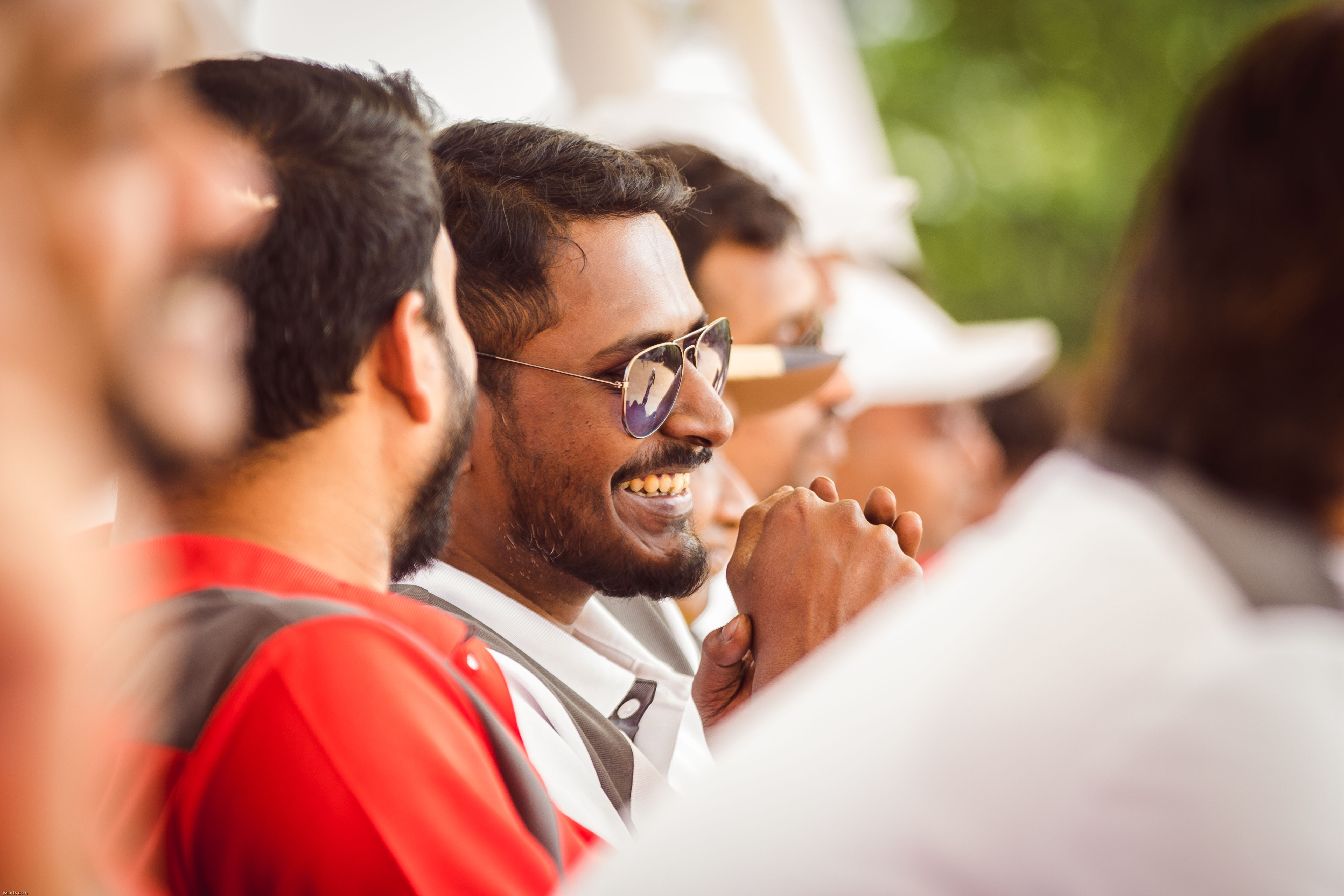A man wearing sunglasses and a beard smiling cheerfully in a lively outdoor crowd.