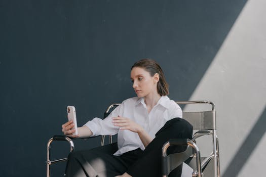 A woman sits in a modern chair using a smartphone against a gray wall.