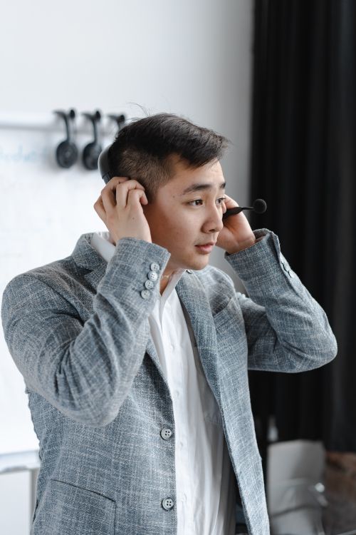 Free Asian man in a gray suit adjusting his headset for a call. Ideal for customer service or business themes. Stock Photo