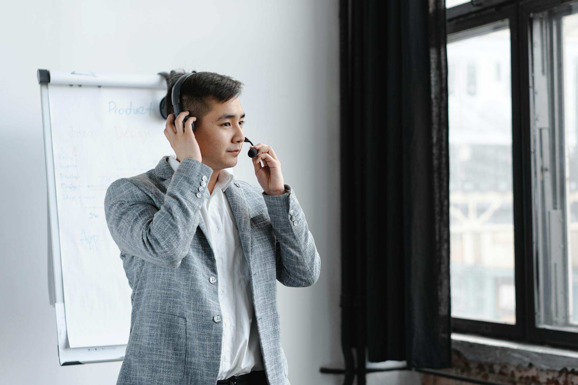 Asian call center agent wearing a headset, handling customer service at an office with a window view.