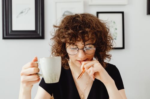 Free A woman with curly hair in glasses holding a mug and pen in a cozy room with framed art. Stock Photo