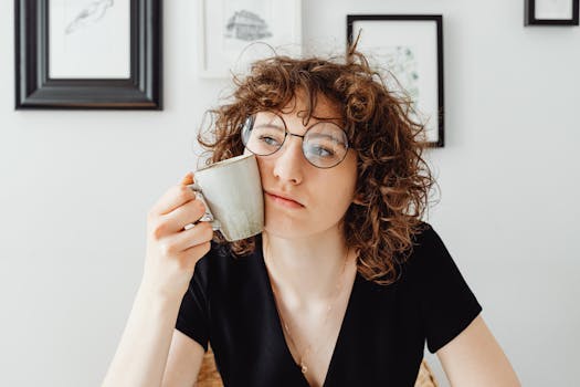 A thoughtful woman with curly hair and glasses holding a coffee mug in a modern room.