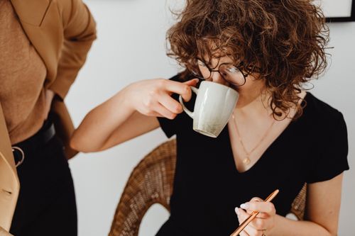 Free A woman with curly hair drinks coffee in a cozy indoor setting, wearing glasses and a black top. Stock Photo