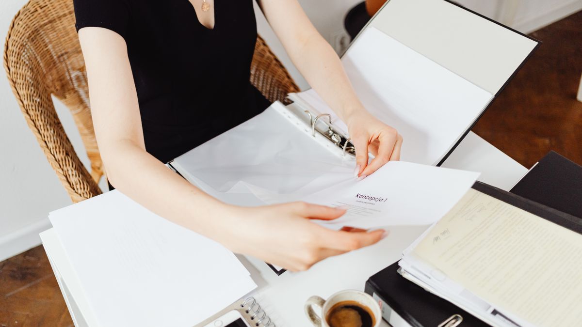 A woman seated at a desk organizing documents in an office setting with coffee and smartphone nearby.