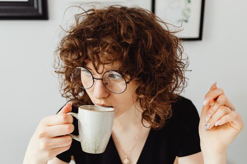Free Caucasian woman with curly hair and glasses enjoying coffee indoors. Stock Photo