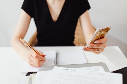 Woman in office multitasking with smartphone and notes, focusing on productivity.