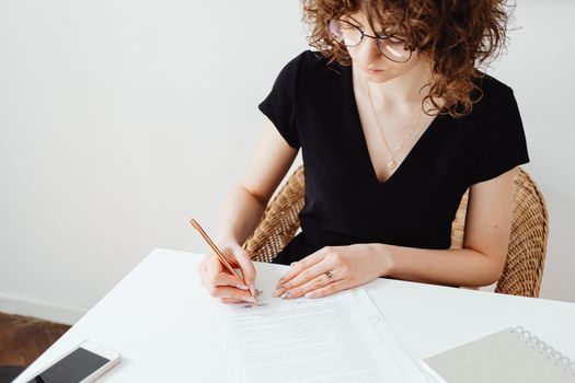A woman in glasses and black top writing on documents at a white table, focusing intently.