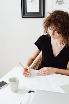 A young woman with curly hair writes at a desk in a modern setting. Perfect for work and lifestyle themes.