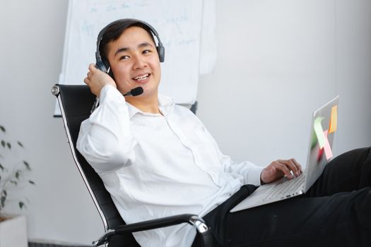 Smiling young man in white shirt using laptop with headset in office setting.