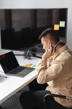 Man in beige jacket using a headset and laptop in a modern office with a large monitor.