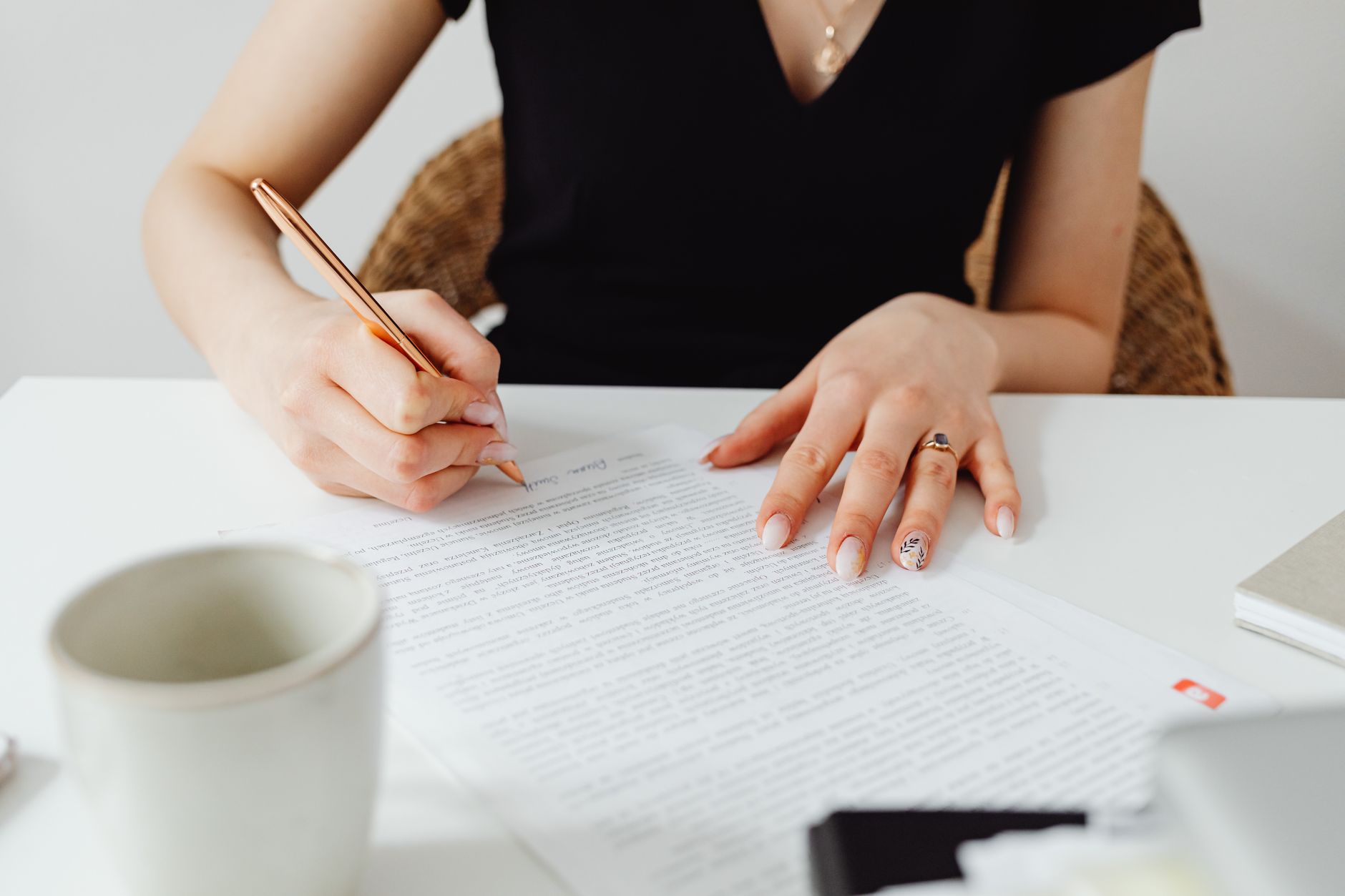 Close-up of a woman writing on papers at a desk with a cup nearby.