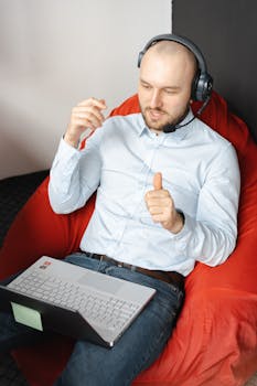 Caucasian man in white shirt using laptop with headset on beanbag chair, engaging in remote work.