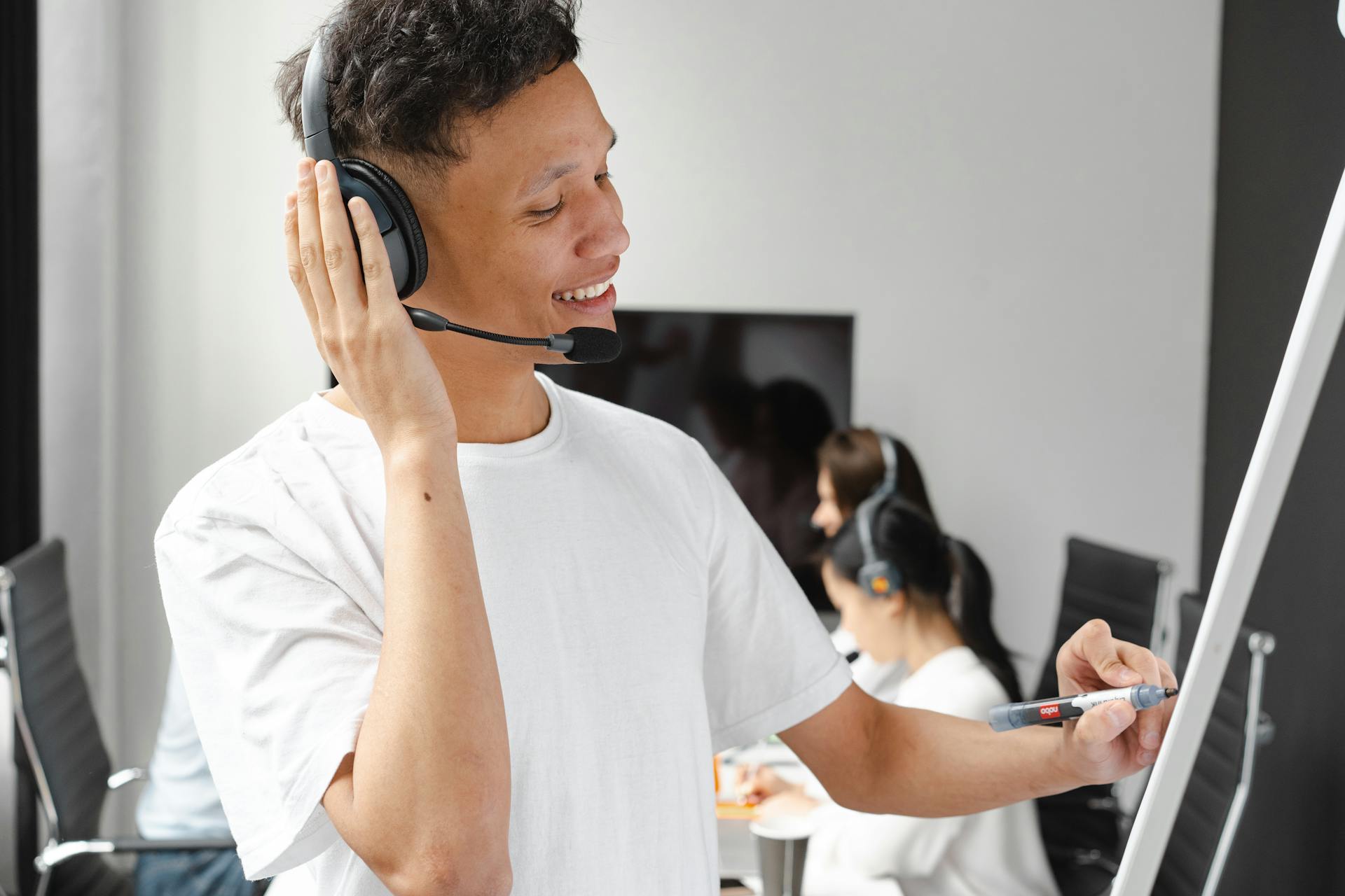 A cheerful customer service agent with headphones writing on a whiteboard in an office setting.