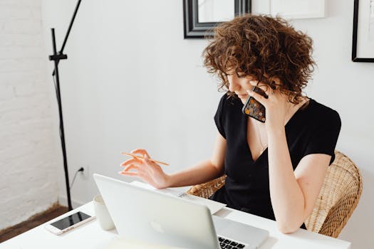 Woman sitting at table, talking on phone while working on laptop in a home office setting.