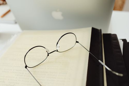 Free Round eyeglasses resting on an open book beside a laptop in a quiet study atmosphere. Stock Photo