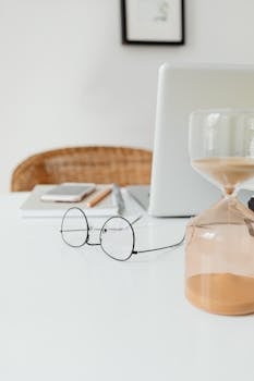 A clean and modern workspace featuring an hourglass and eyeglasses on a desk.
