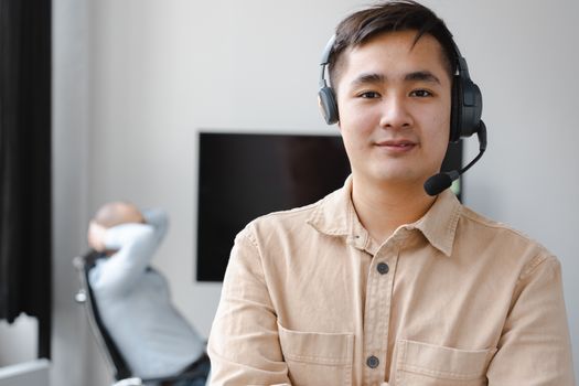 Asian man wearing a brown shirt and headset smiles in a modern office environment.