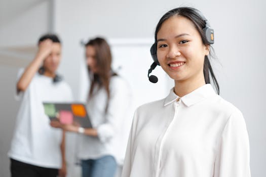 Young woman in a call center with colleagues in the background, smiling and wearing headphones.