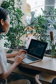 Businesswoman checks financial graphs on laptop in cozy café, surrounded by plants, showcasing remote work lifestyle.