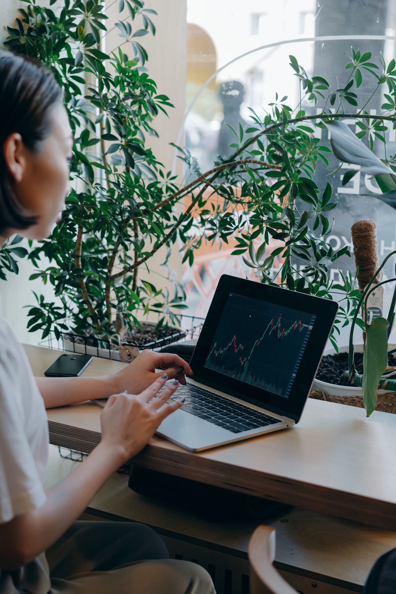 Free Businesswoman checks financial graphs on laptop in cozy café, surrounded by plants, showcasing remote work lifestyle. Stock Photo