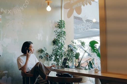 Asian woman multitasking with phone and laptop in a cozy café environment.