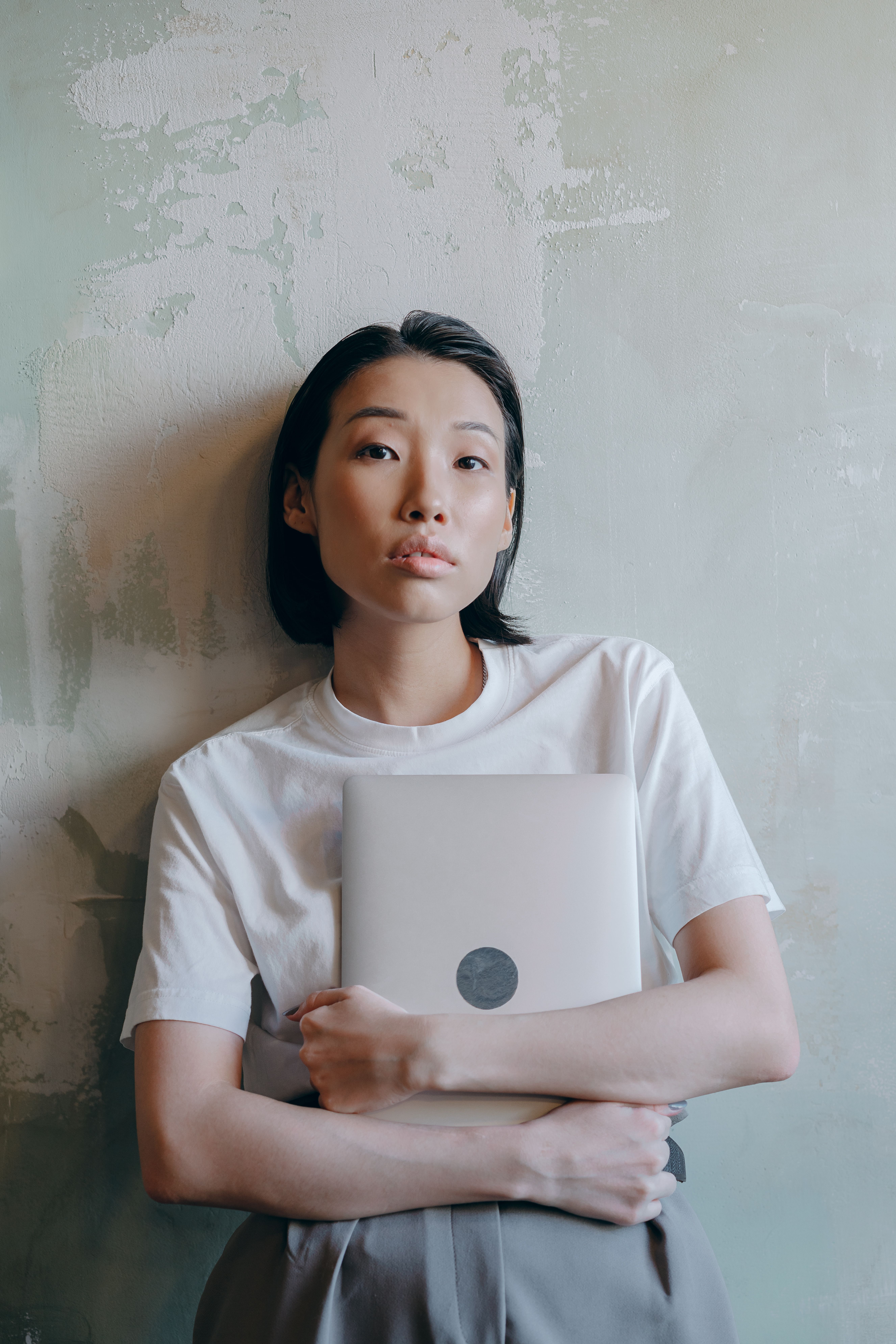 Asian woman in a white shirt holding a laptop, leaning against a textured wall.